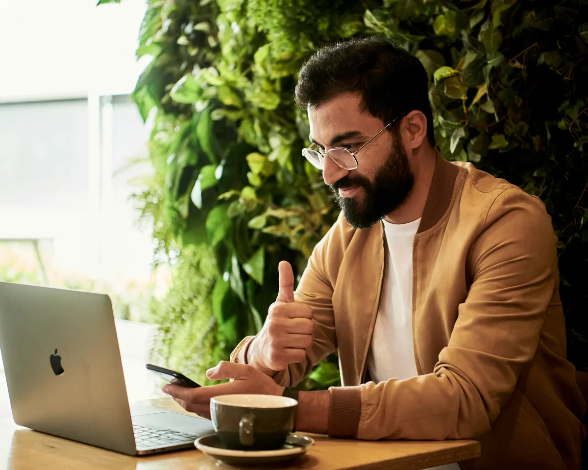 A man at a cafe table smiling at his laptop and giving it a thumbs up, a phone in his other hand and a plant wall behind him