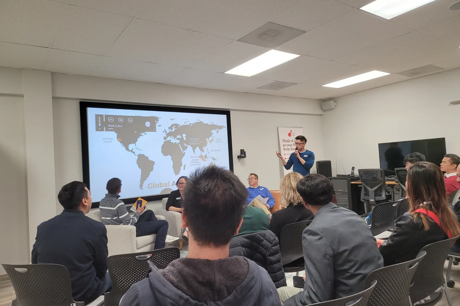 A room of people seated on chairs and sofas facing a screen that shows a world map, a speaker standing at the right