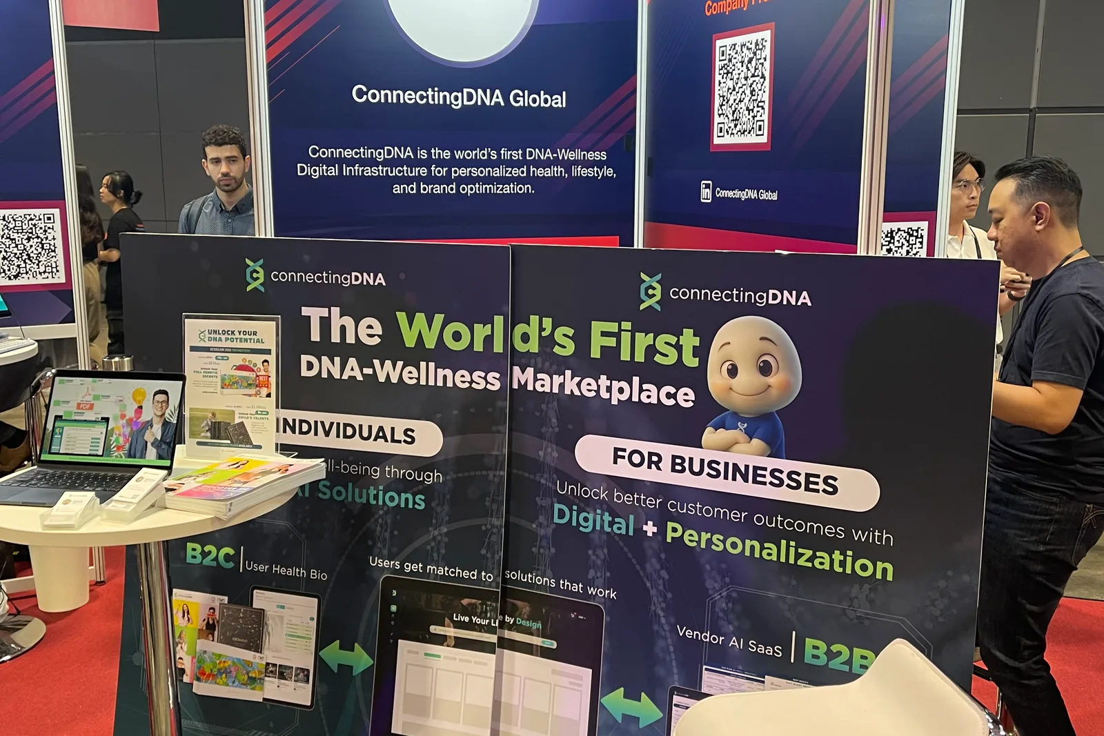 The ConnectingDNA booth at a trade show, its backdrop reading The World's First DNA-Wellness Marketplace, with a laptop on a stand and visitors at the side
