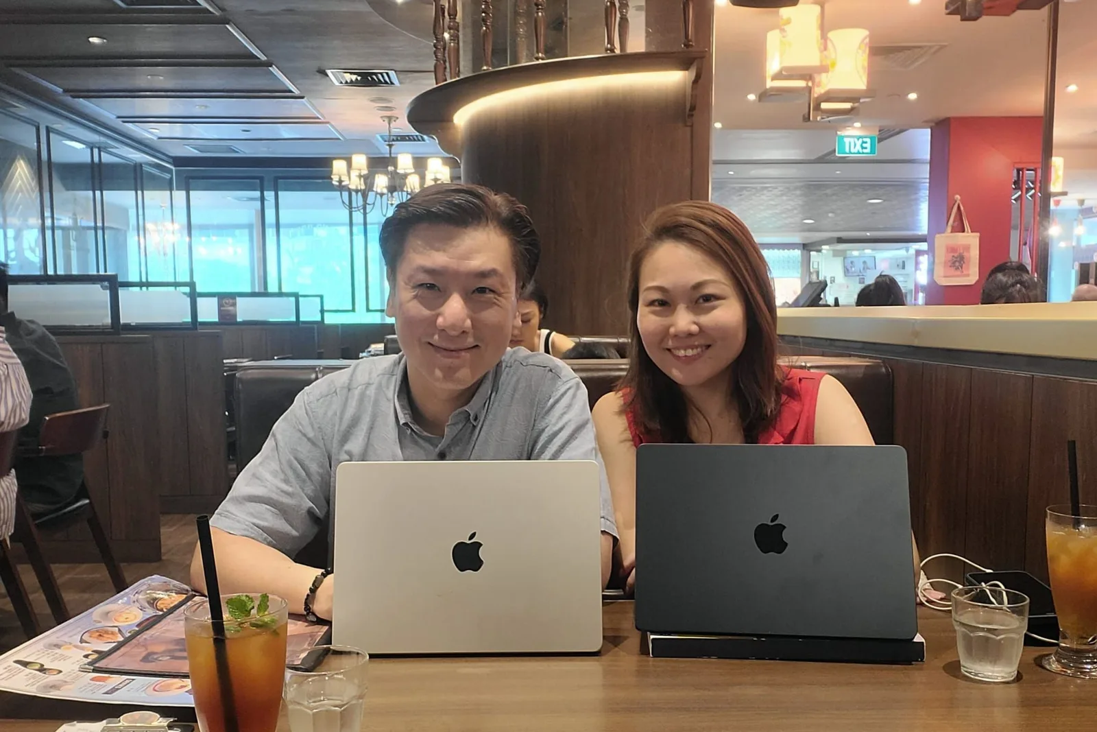 Cedric Lee in a grey shirt and Choo Ee Ting in a red sleeveless top, smiling side by side behind their laptops at a restaurant table, iced drinks in front of them