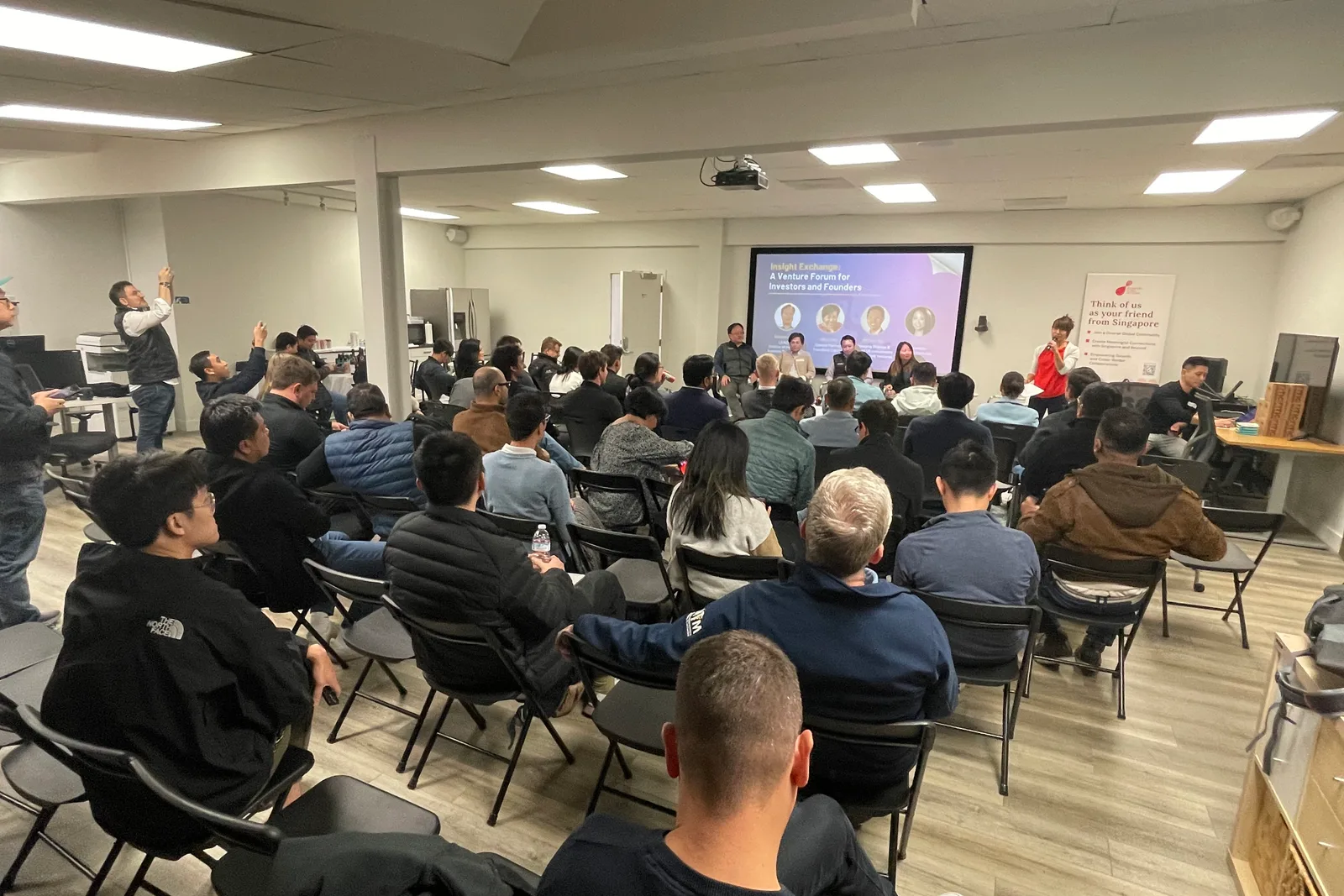 A full room seen from the back rows, a panel of four seated under a screen reading Insight Exchange: A Venture Forum for Investors and Founders, a banner at the right reading Think of us as your friend from Singapore
