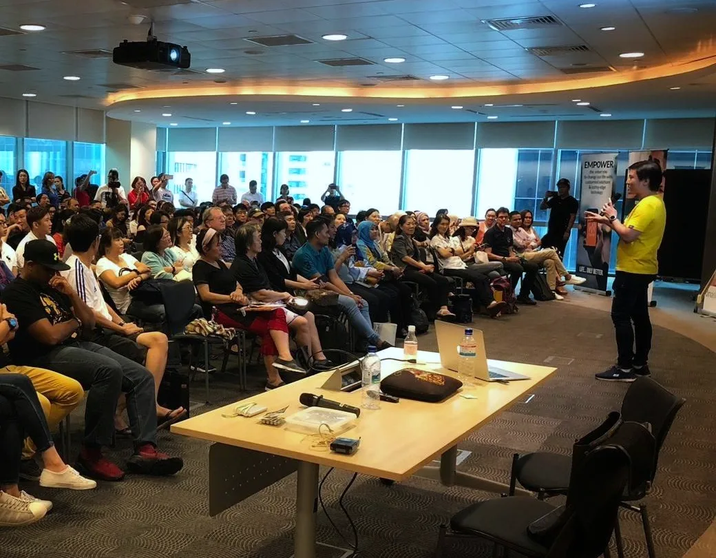 Cedric Lee in a yellow T-shirt speaking into a microphone to a packed room, rows of seated attendees and more standing along the windows at the back