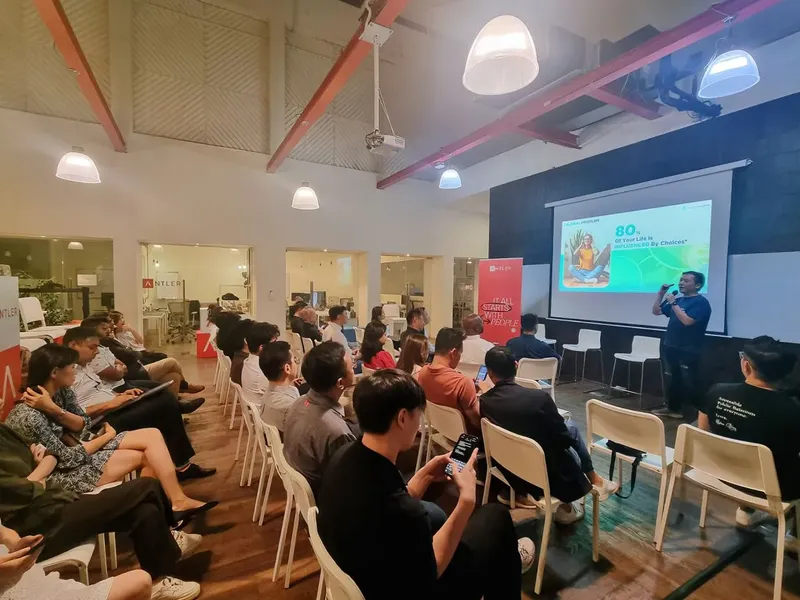 Rows of people in white chairs under a high ceiling with red beams, facing Cedric Lee beside a projected slide