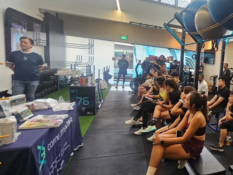 Cedric Lee standing beside a ConnectingDNA table as gym members sit on benches and the floor between squat racks and coloured weight plates