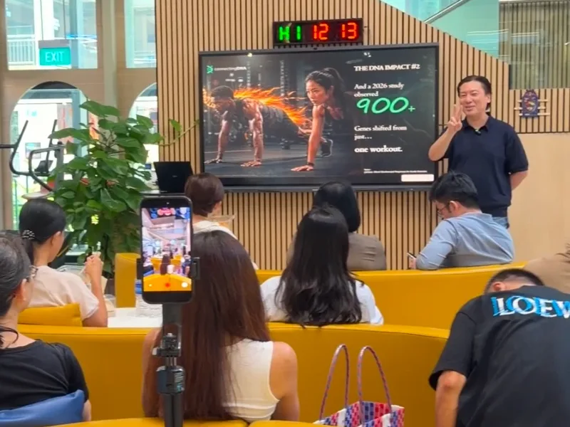 Cedric Lee raising two fingers beside a screen reading 900+ genes shifted from just one workout, an audience on yellow sofas, one filming him on a phone