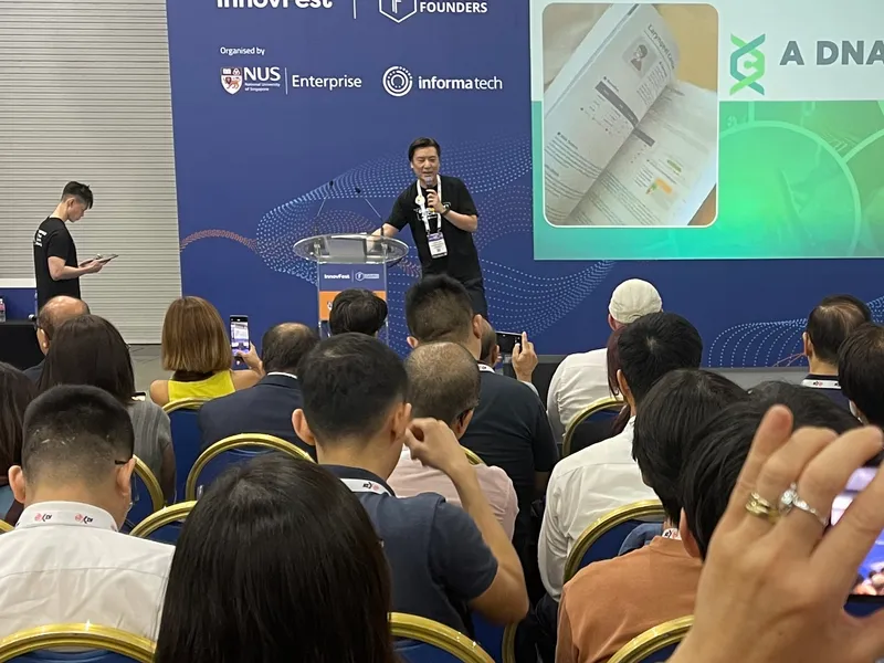 Cedric Lee on the InnovFest stage beside a slide reading A DNA Report, rows of seated attendees, one filming him on a phone