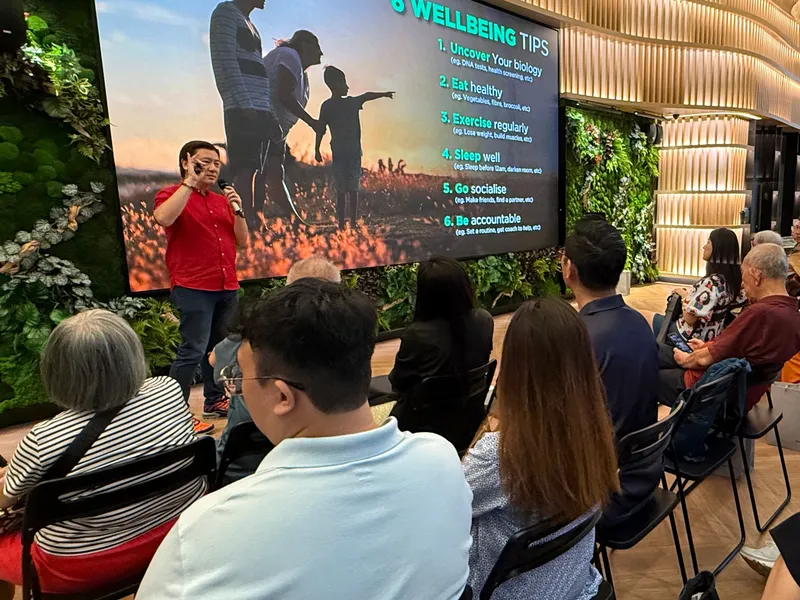 Cedric Lee in a red shirt, a microphone in hand, beside a screen of six wellbeing tips, an audience seated in rows under a curved wooden ceiling