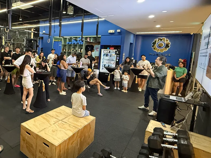 Cedric Lee talking by a wall of screens at Mobilus, a standing crowd around high tables and a child seated on wooden plyometric boxes