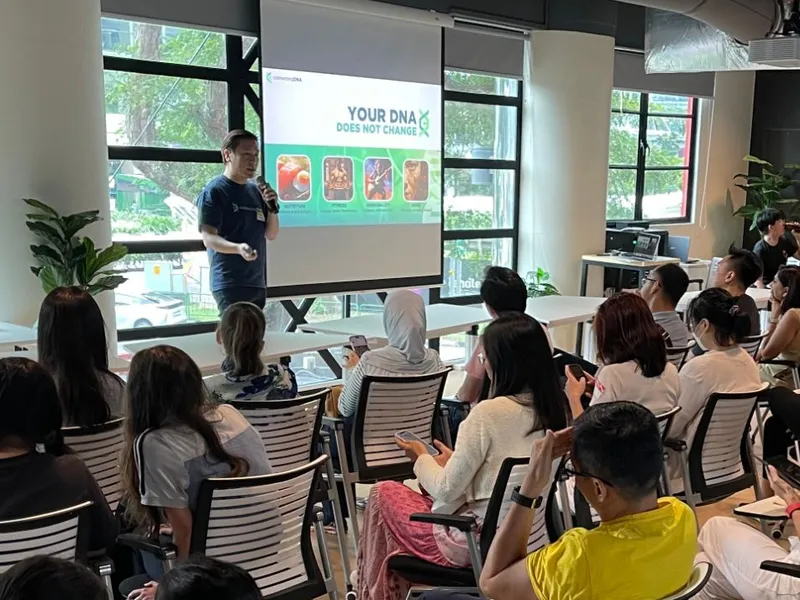 Cedric Lee with a microphone beside a screen reading Your DNA Does Not Change, adults and children seated in rows of white chairs