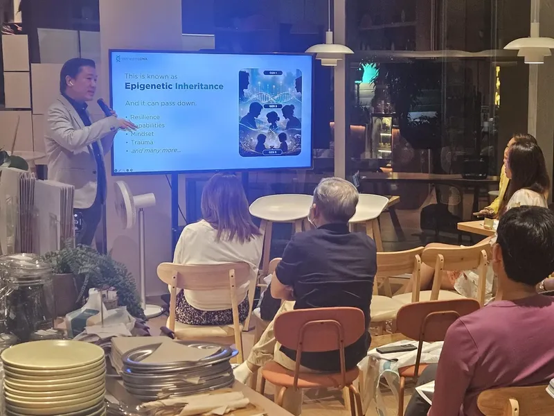 Cedric Lee presenting beside a screen reading This is known as Epigenetic Inheritance, parents seated at cafe tables