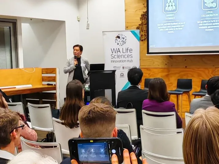Cedric Lee with a microphone beside a WA Life Sciences Innovation Hub banner and a projected slide, an audience in white chairs, one filming on a phone