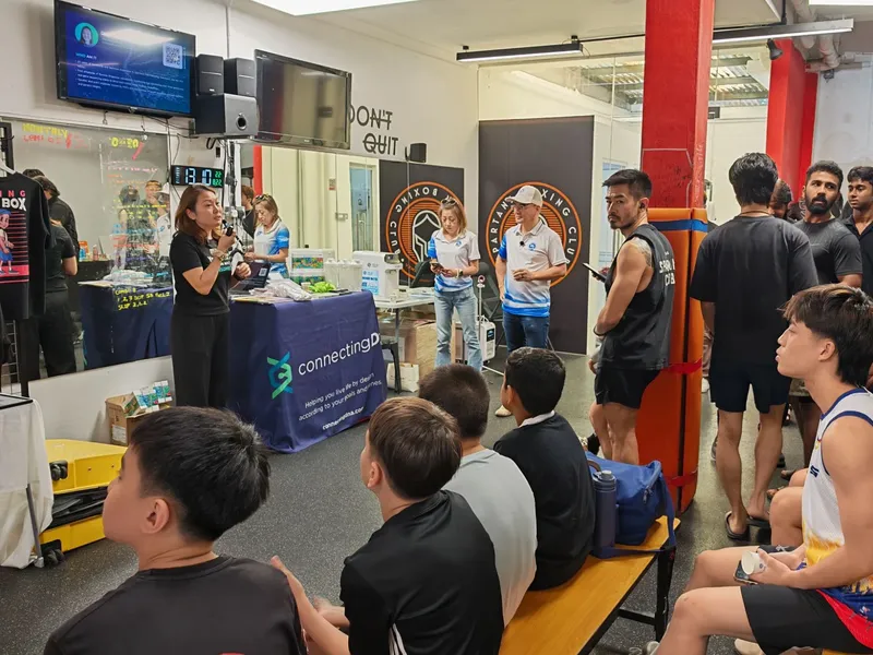 A speaker with a microphone beside a ConnectingDNA table at Spartans Boxing Club, young boxers on benches and a standing crowd by a red pillar