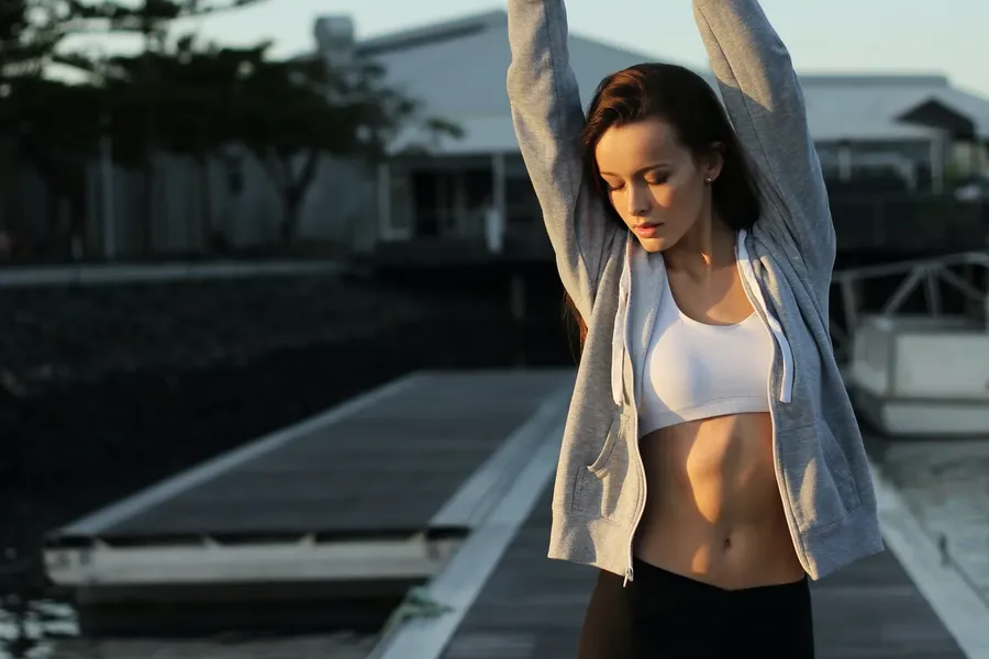 A woman in a grey hoodie and sports top stretching her arms overhead on a wooden dock in early light