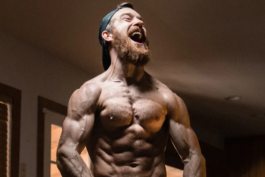 A bearded, heavily muscled man, shirtless and laughing with his head thrown back