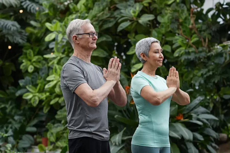 An older man and woman in sportswear standing in a garden, palms pressed together at the chest