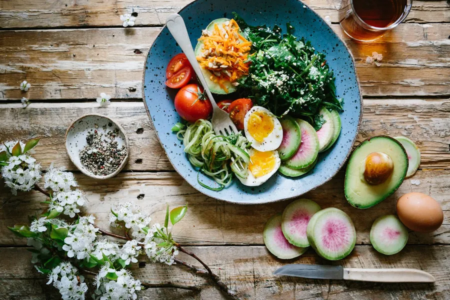 A speckled blue bowl of greens, cherry tomatoes, grated carrot, spiralised cucumber, radish and a halved boiled egg on a wooden table, half an avocado and a sprig of white blossom beside it