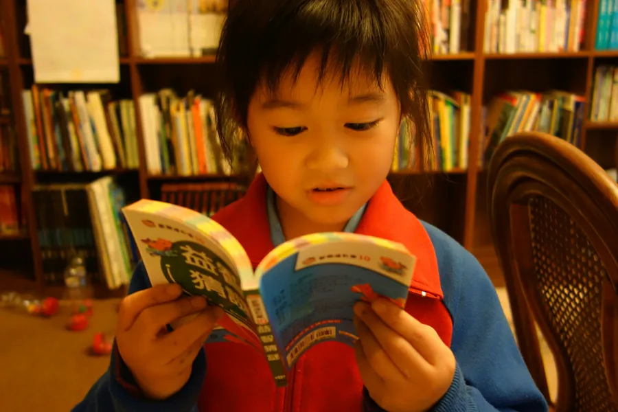 A young child in a red vest reading a picture book aloud in front of shelves full of books