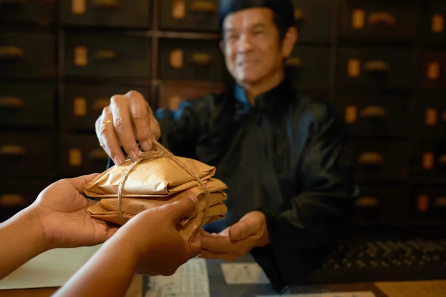 A smiling older herbalist in a dark tunic hands a stack of paper-wrapped herb parcels tied with string across a counter, wooden medicine drawers behind him